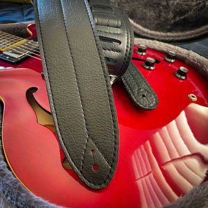 3-inch wide black leather guitar strap with stitch down the middle on top of a red Gibson 335 guitar